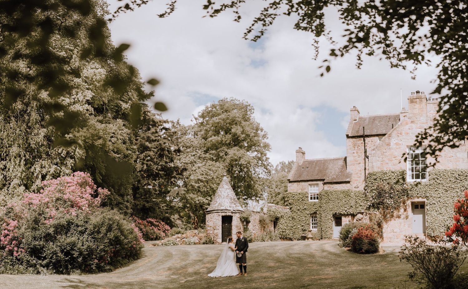 Wide shot of wedding couple on the grounds of a stone castle estate with pink flowers.