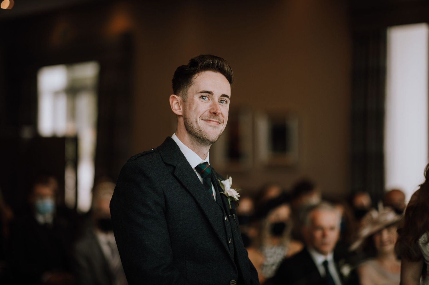 A groom in a dark suit and boutonniere stands during wedding ceremony.