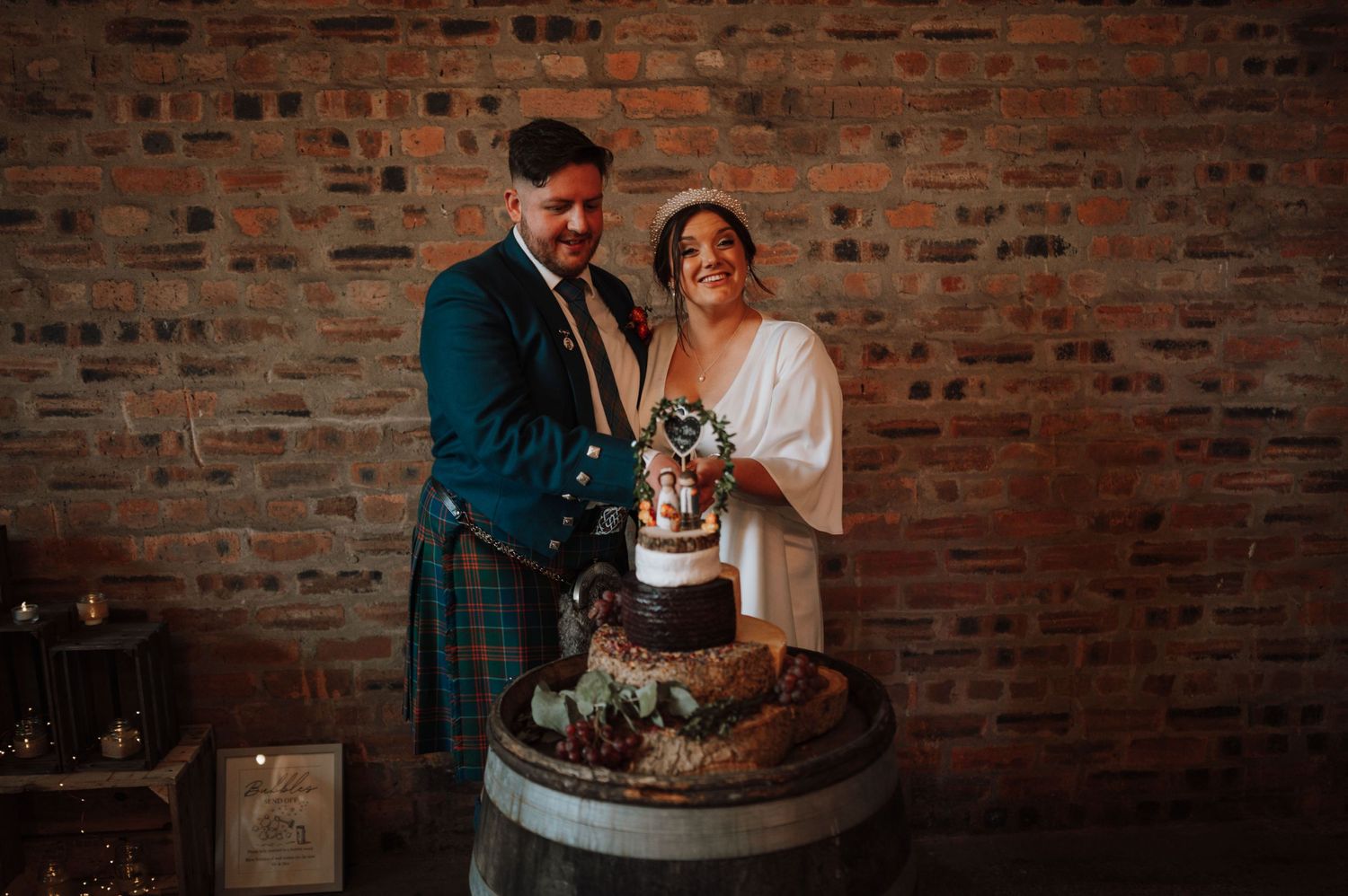Wedding couple cuts cake on rustic wooden barrel against brick wall background.