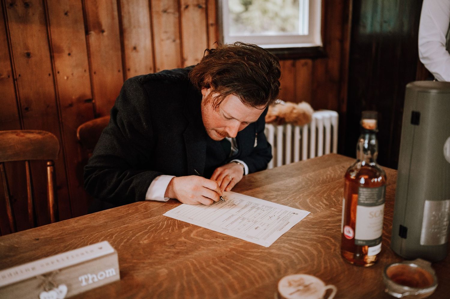 Person leaning over wedding paperwork at rustic wooden table with bottle nearby.