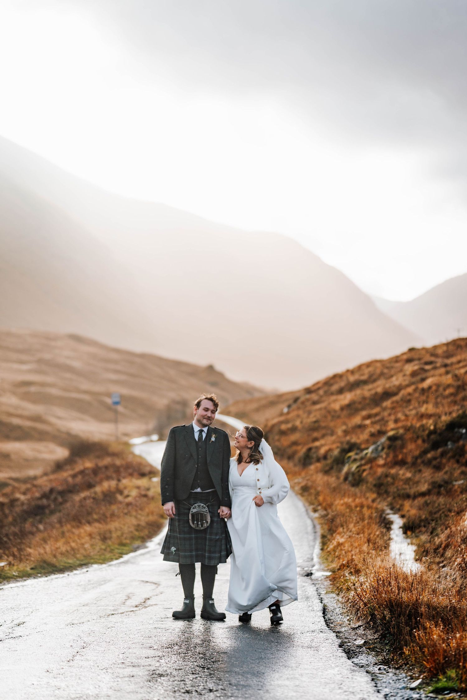 A couple in wedding attire walking on a wet road through misty Scottish Highlands with mountains in background.