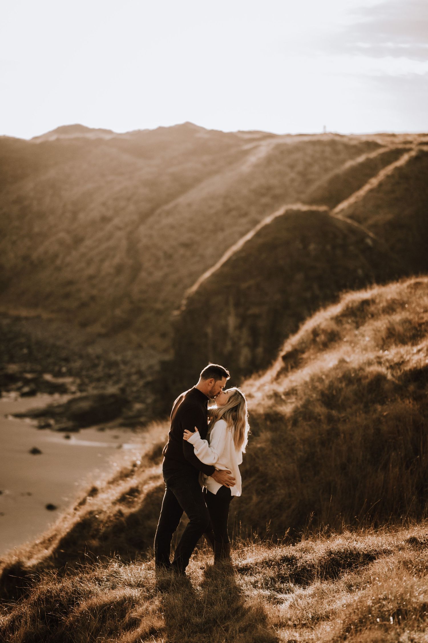 A couple embraces on a scenic hillside overlooking a dramatic coastline at sunset.