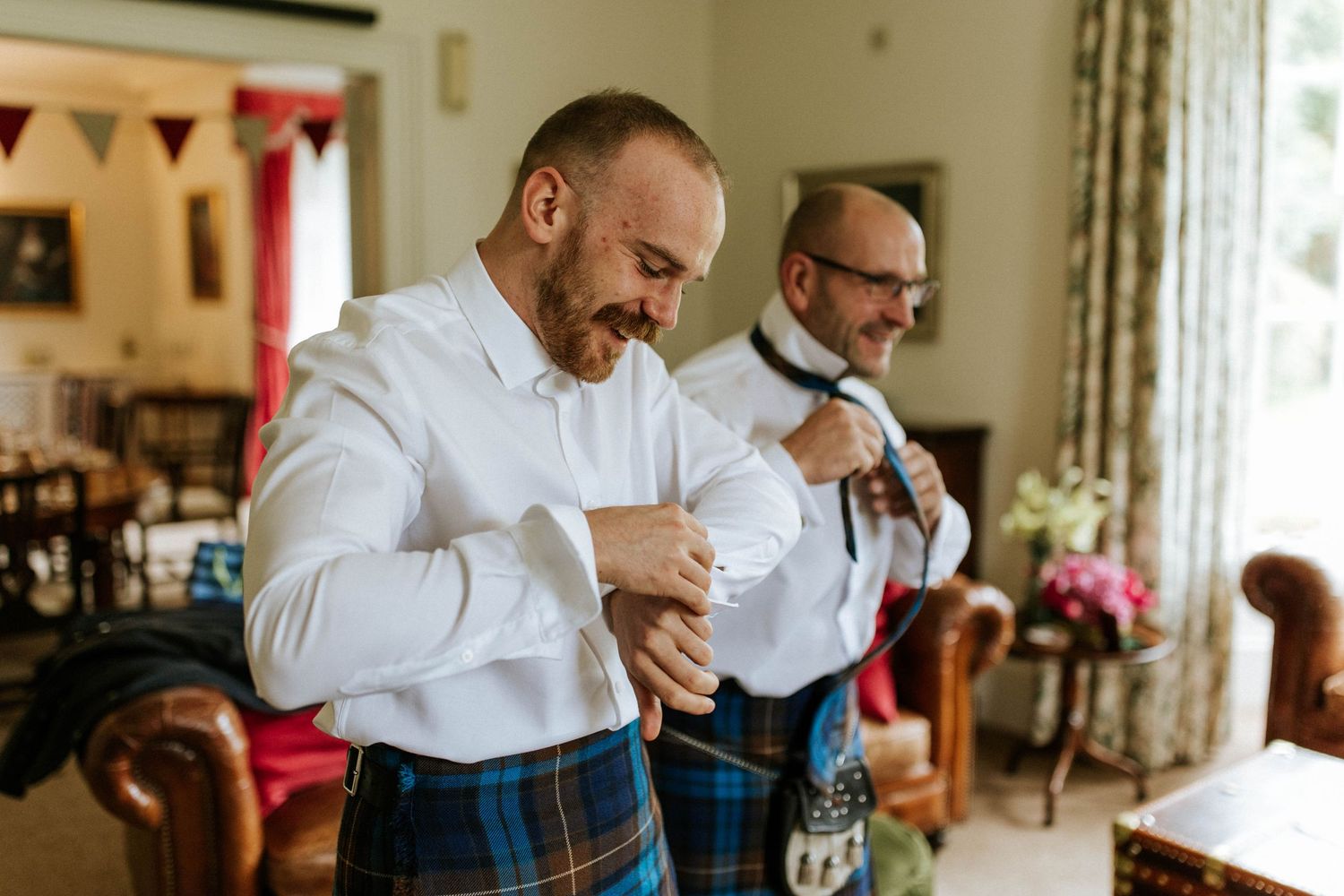 Groomsmen in traditional Scottish kilts sharing drinks and laughs during wedding preparations.