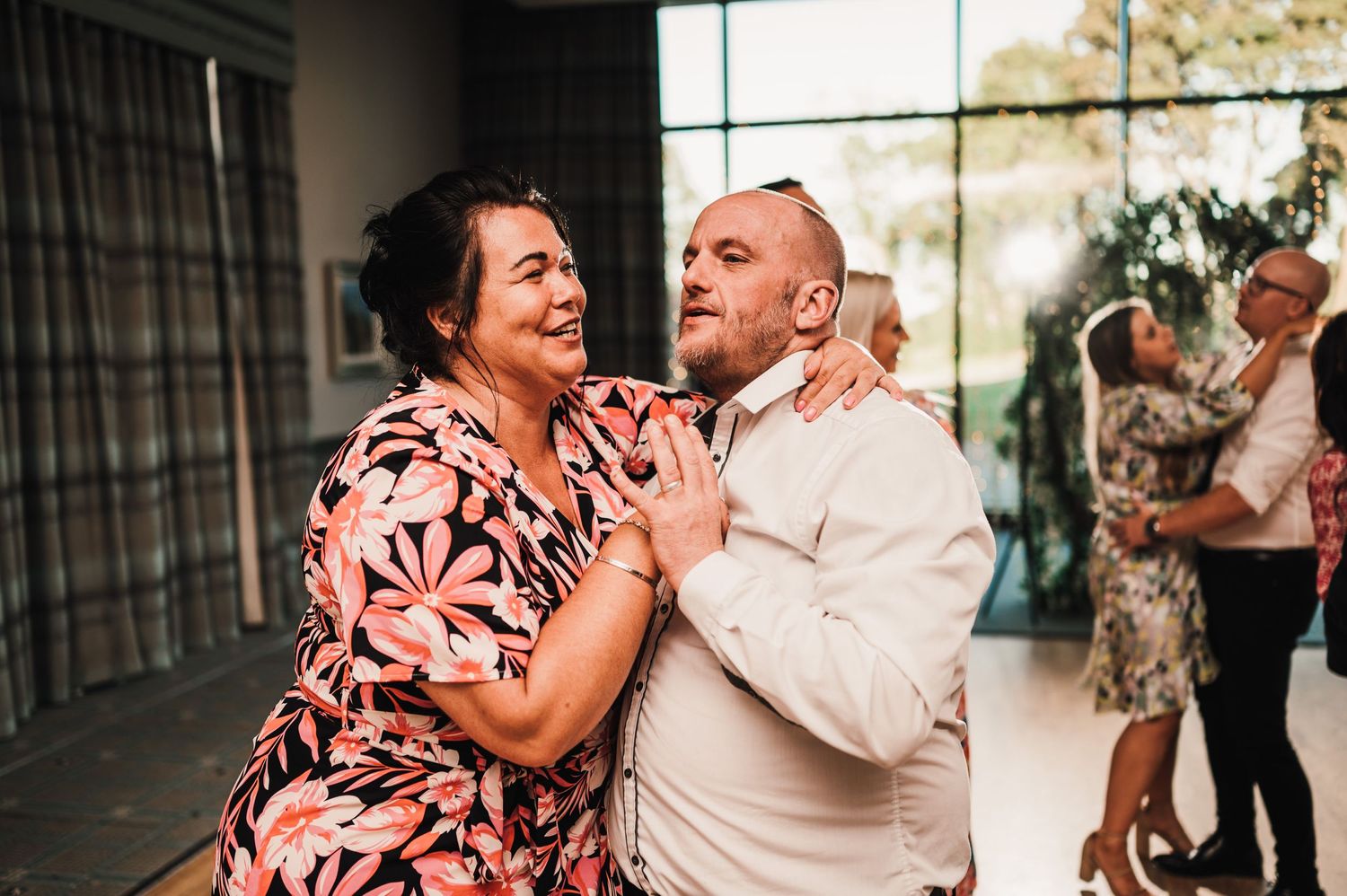 A couple shares a joyful moment dancing together at a wedding reception with guests in the background.