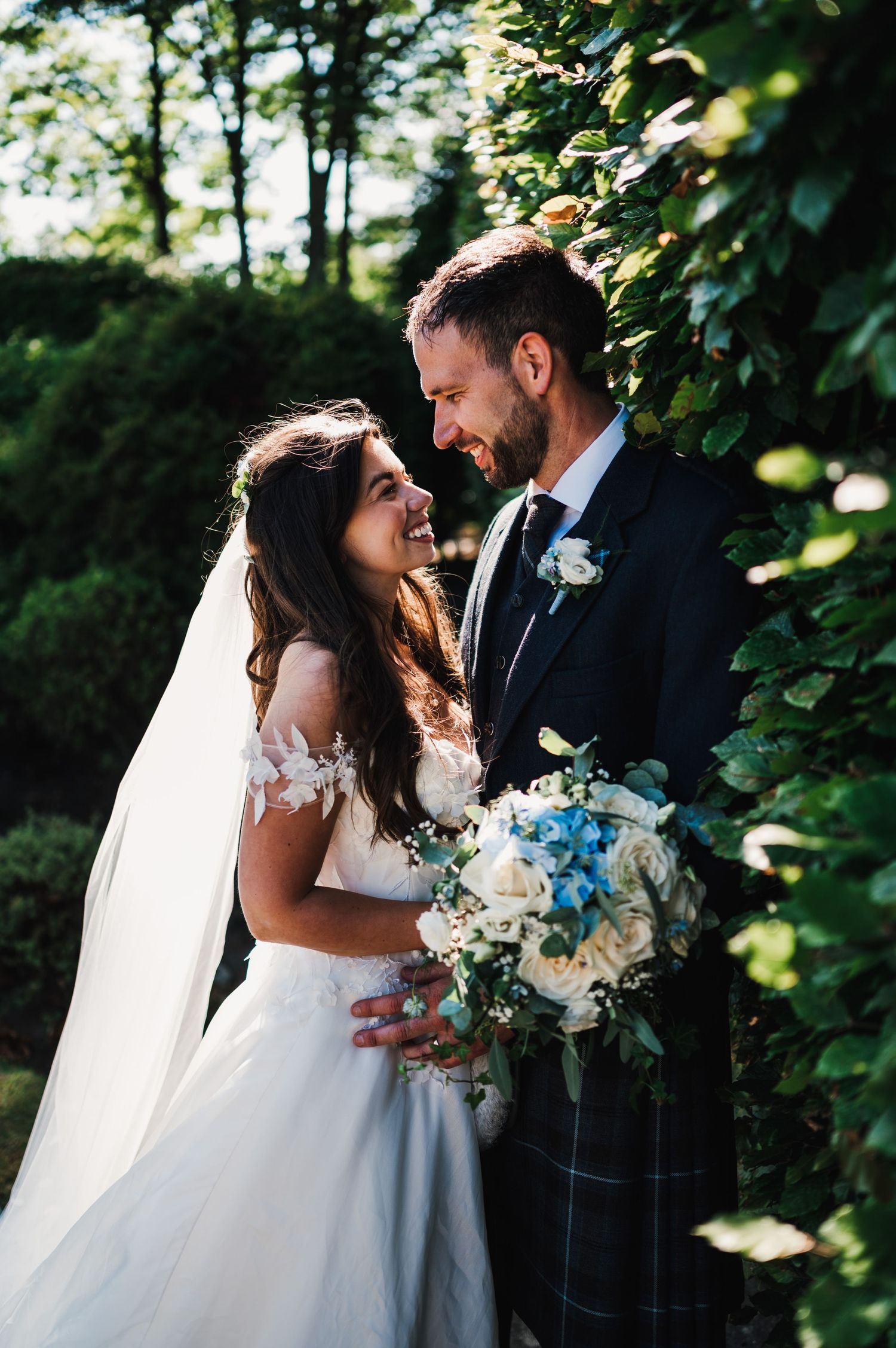 A tender moment captured between newlyweds among garden greenery with blue and white floral bouquet.
