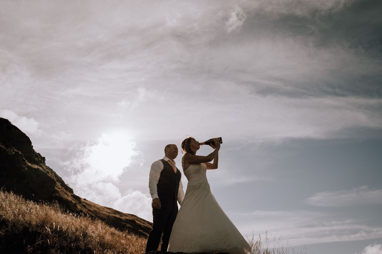 Silhouettes of newlyweds celebrate on a scenic mountain hillside at dusk.