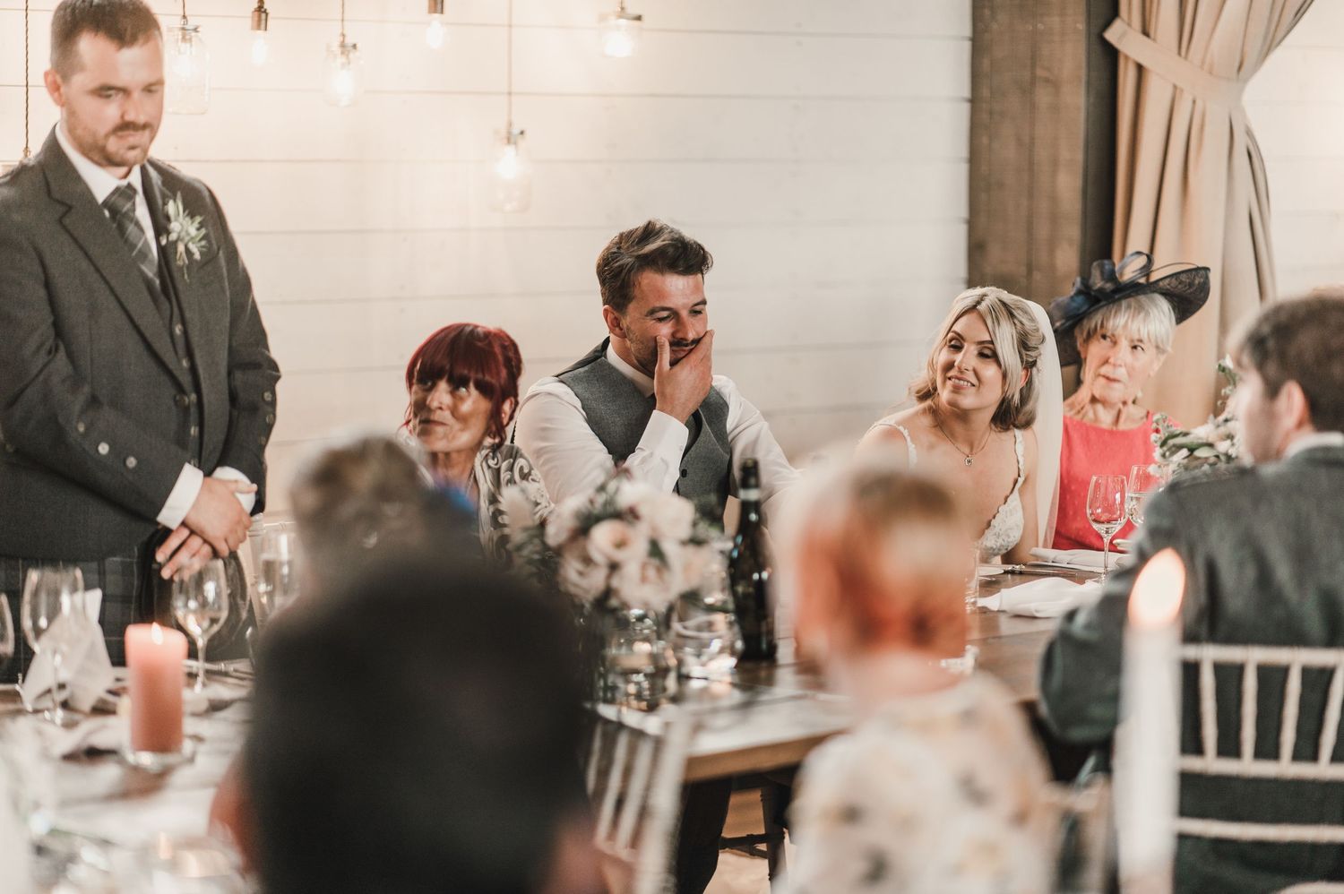 Wedding guests enjoying speeches during a reception dinner with pendant lighting and elegant decor.