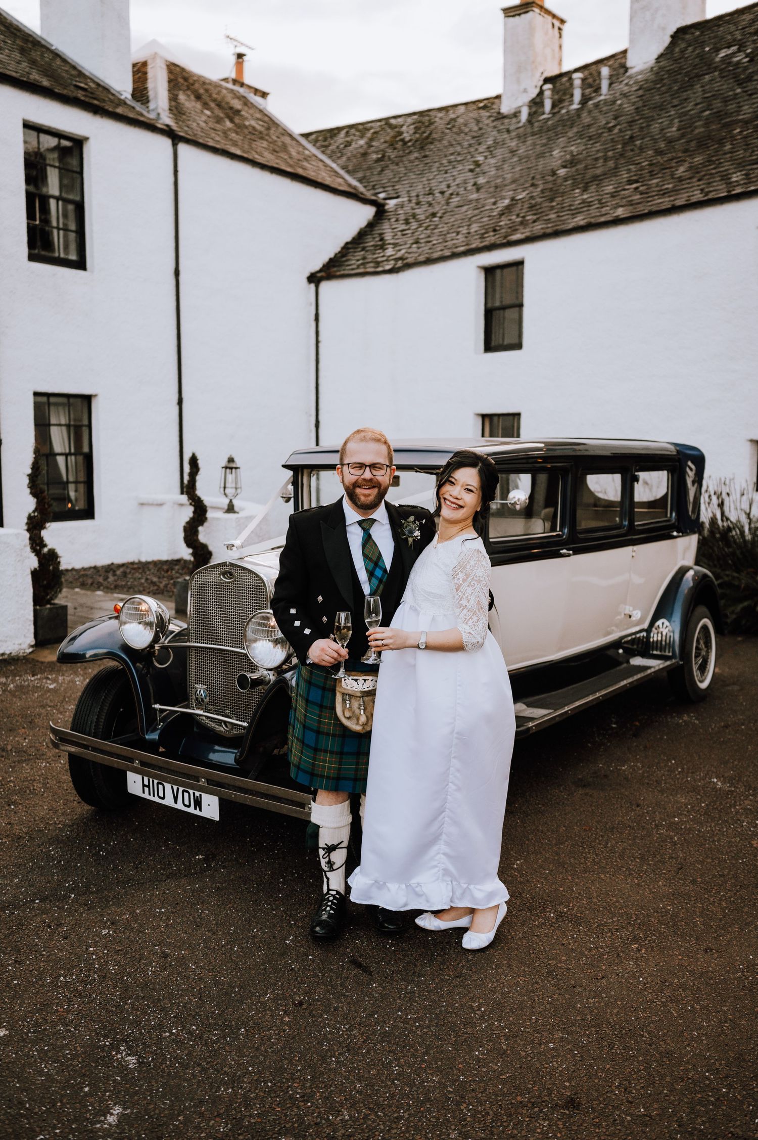 Newlyweds pose by a vintage cream-colored car outside a white Scottish manor house.