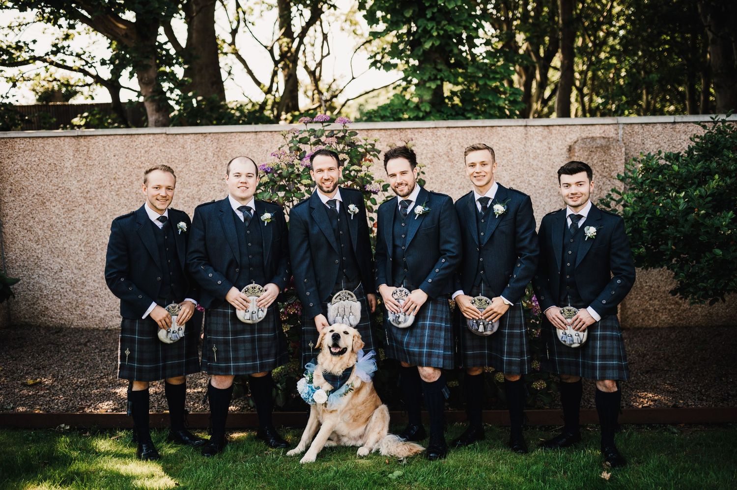 Wedding party wearing traditional Scottish kilts poses with golden retriever on lawn.