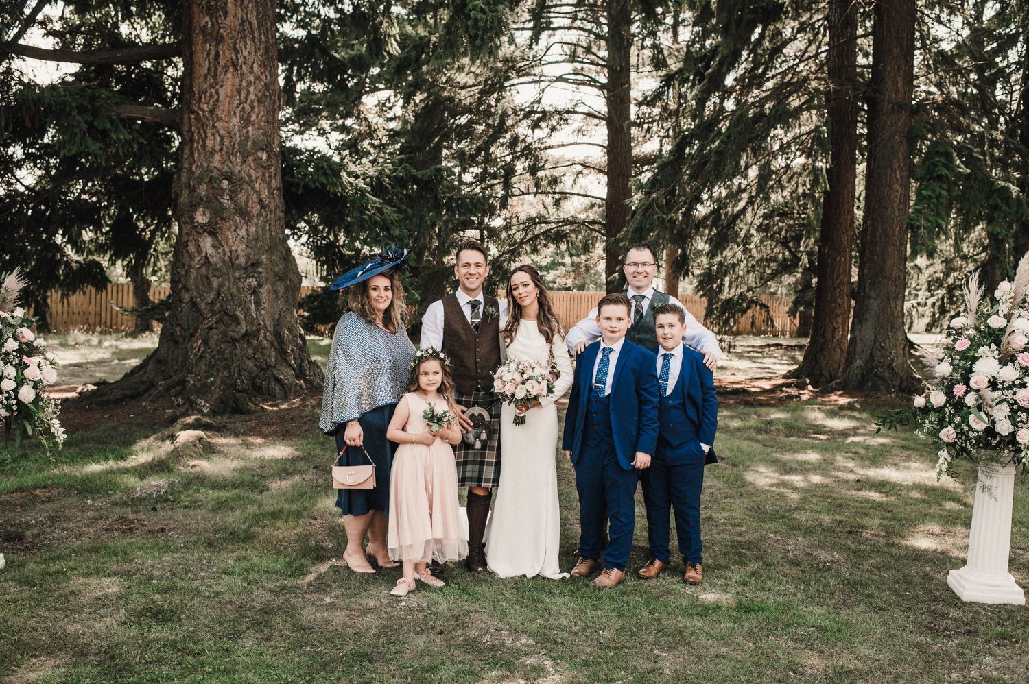 Small wedding party poses together beneath tall pine trees with floral decorations.