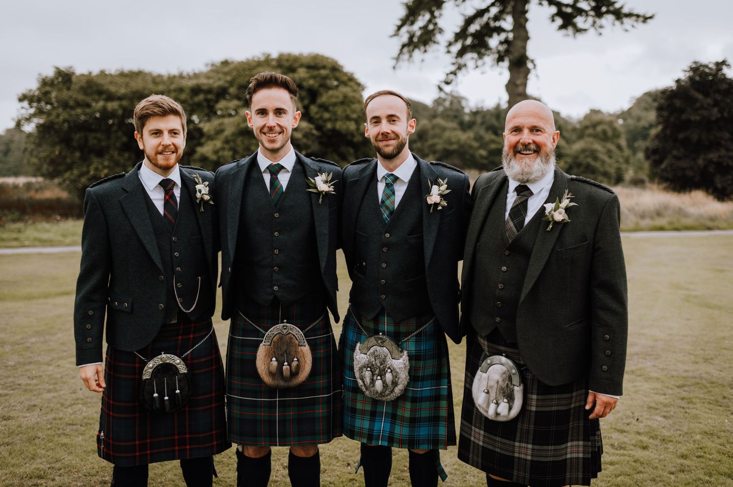 Four men in traditional Scottish kilts and formal wear standing together on grass.