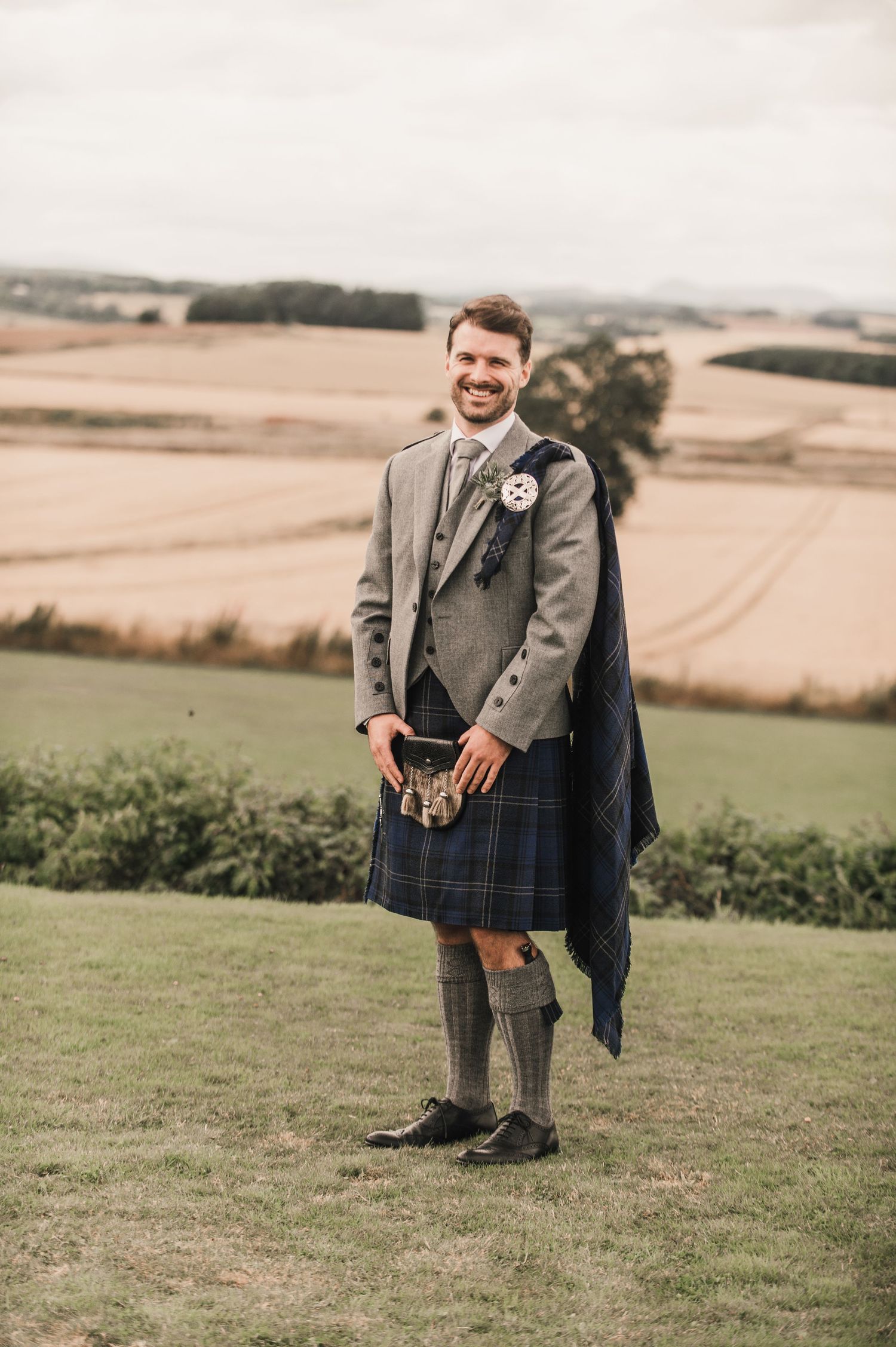 A person wearing a traditional grey Scottish kilt suit stands on a grassy hilltop overlooking rural farmland.