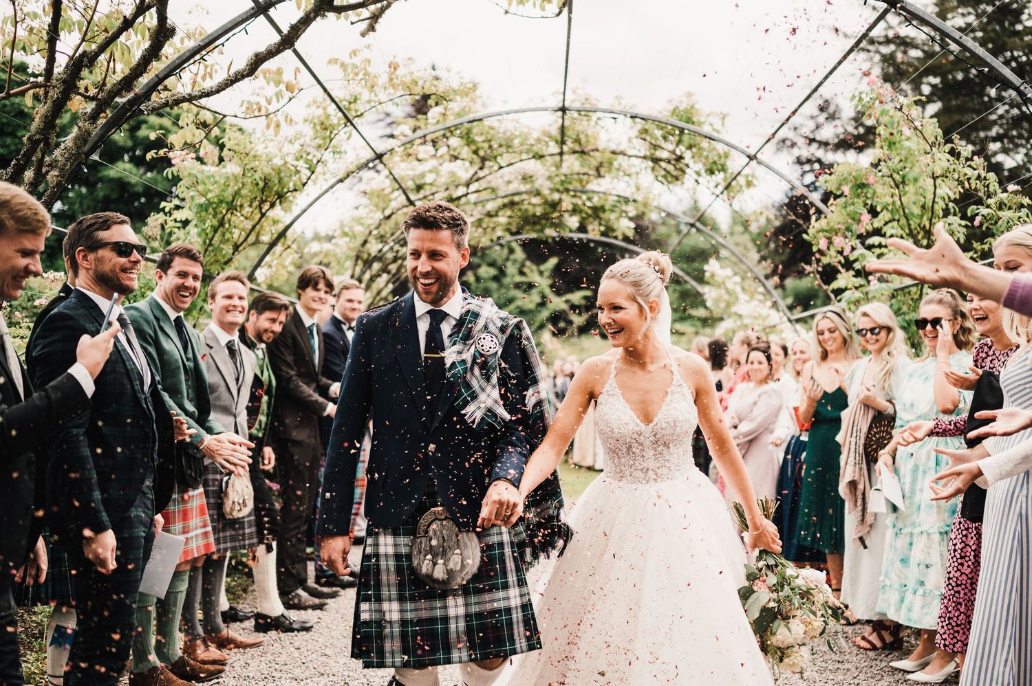 Wedding guests throw confetti as the couple walks through a garden arch tunnel.