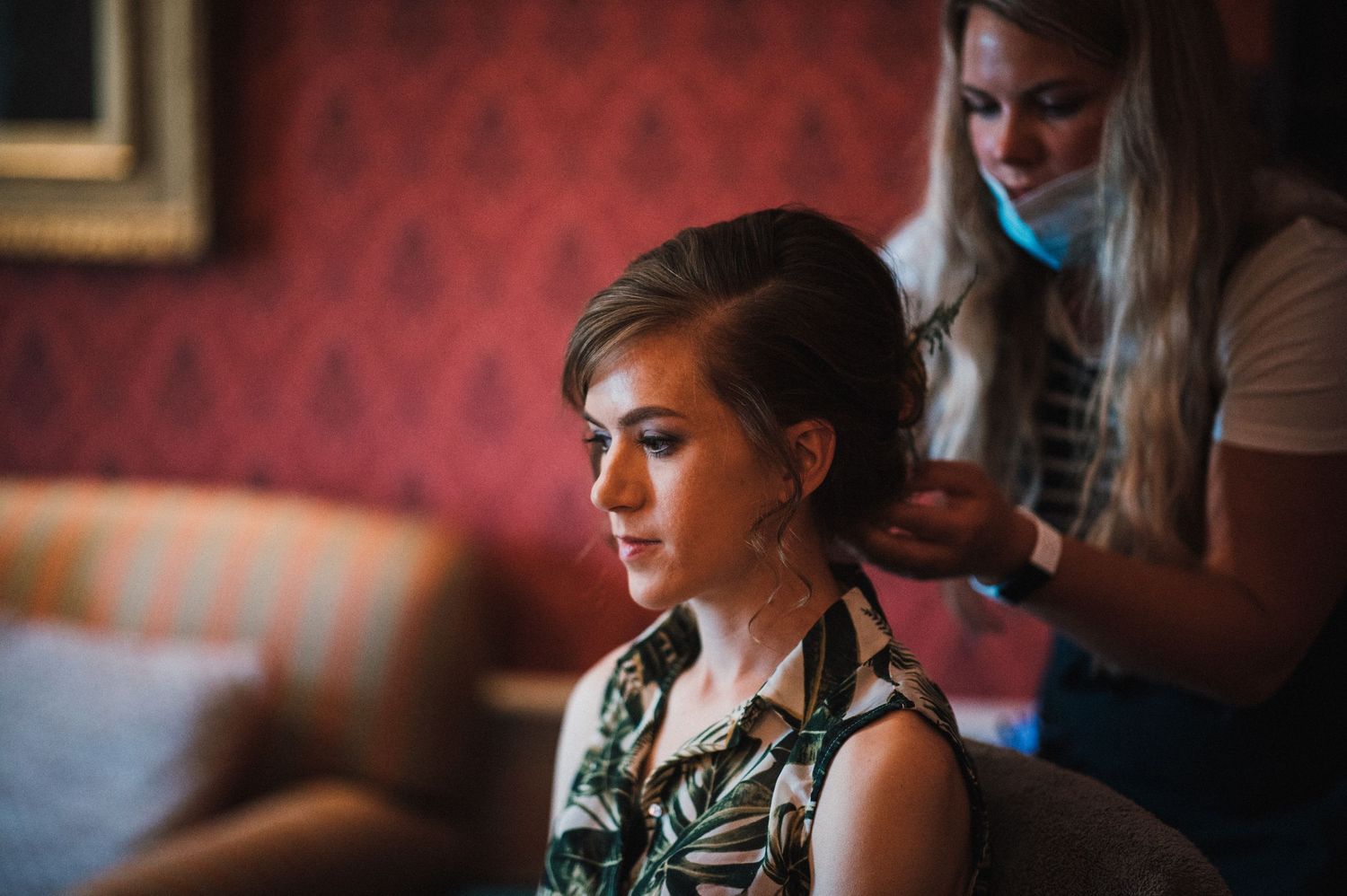 A person getting their hair styled in a room with red wallpaper while wearing a floral print sleeveless top.
