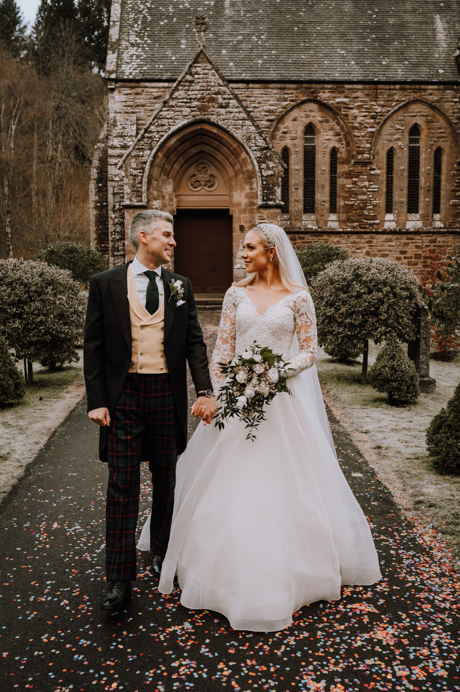 Newlywed couple in elegant attire stands outside gothic stone church surrounded by topiary.