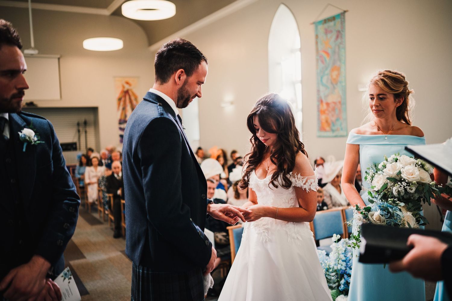 Intimate moment during wedding ring exchange at church ceremony with bridal party looking on.