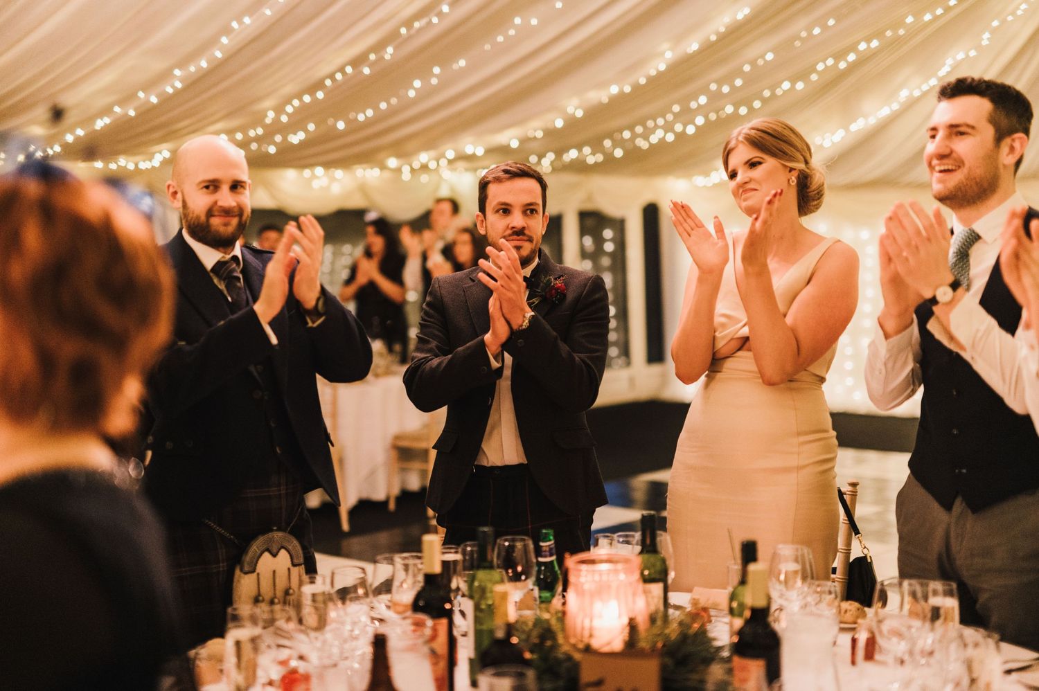 Wedding guests at a reception applauding during an emotional moment under fairy lights.