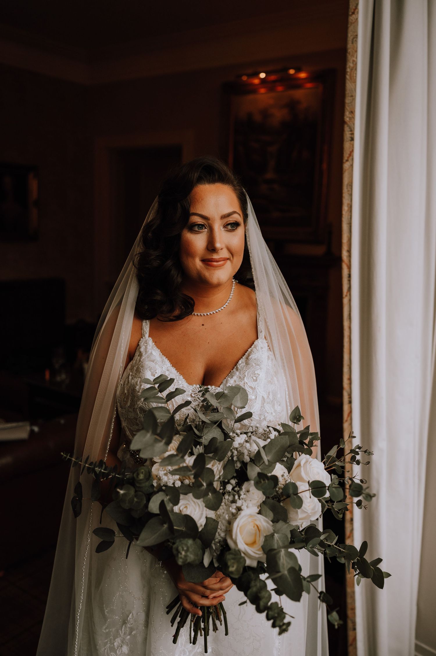 A classic bridal portrait featuring a white dress and eucalyptus bouquet by a window.