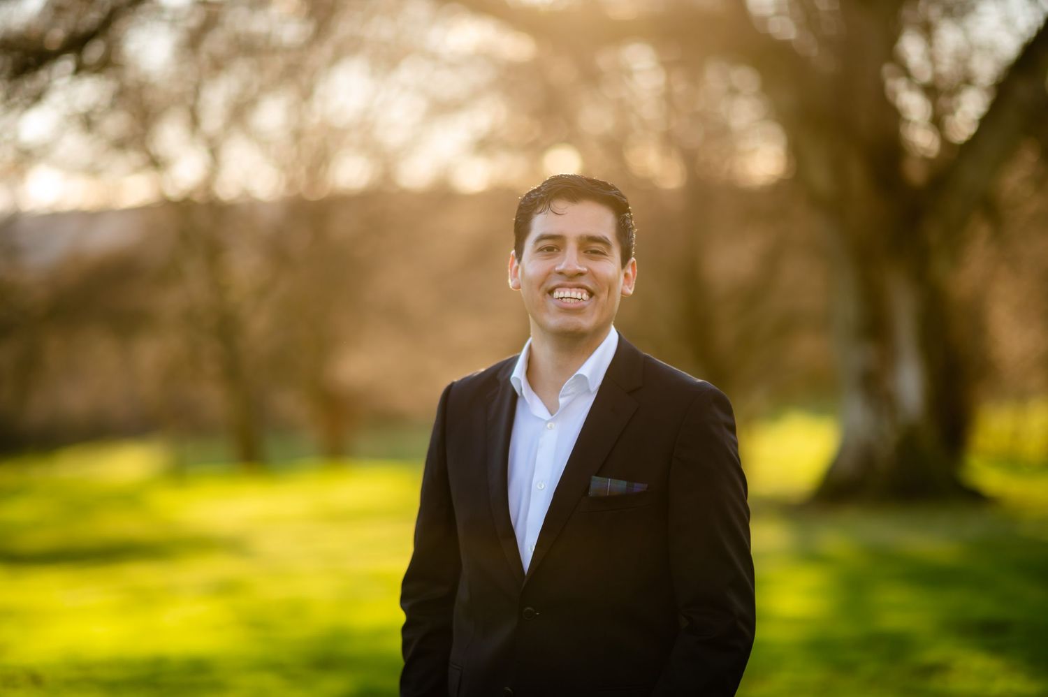 Person in dark suit and white shirt standing outdoors in golden sunlit park setting.