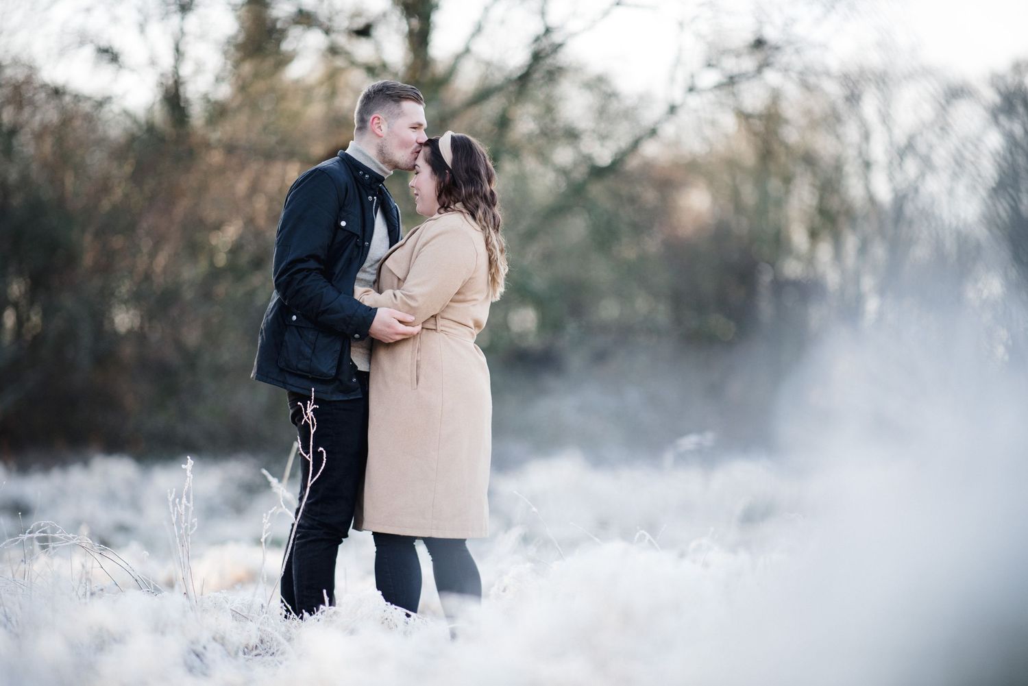 A romantic winter engagement photo with the couple embracing in snowy misty scenery.