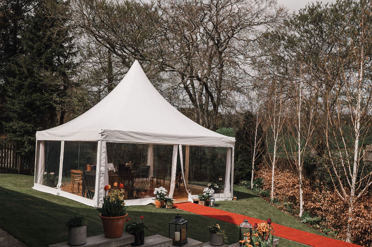 A white wedding tent set up outdoors with a red carpet entrance and surrounding trees.