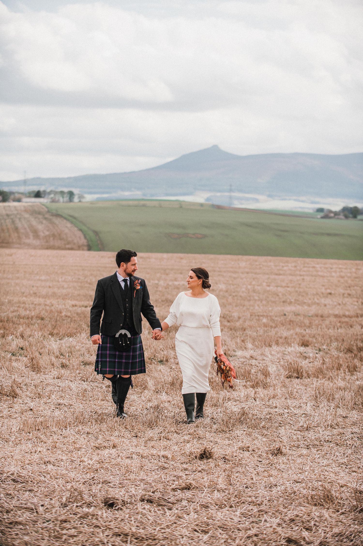 A couple holding hands walks through a rural field with rolling hills and mountains in the background.