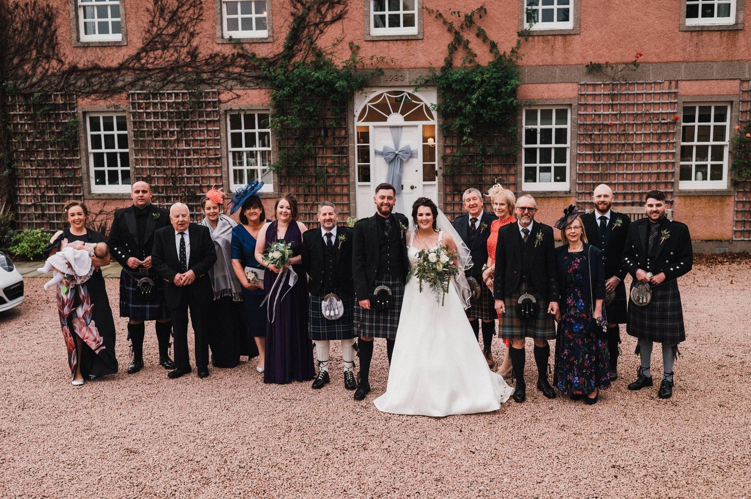 Wedding party in traditional Scottish attire gathered outside historic brick building on gravel path.