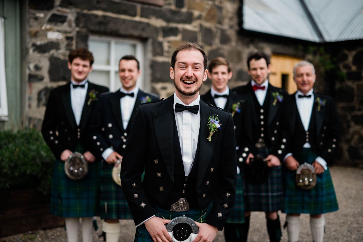 Wedding party in matching kilts and formal attire smiling outdoors.