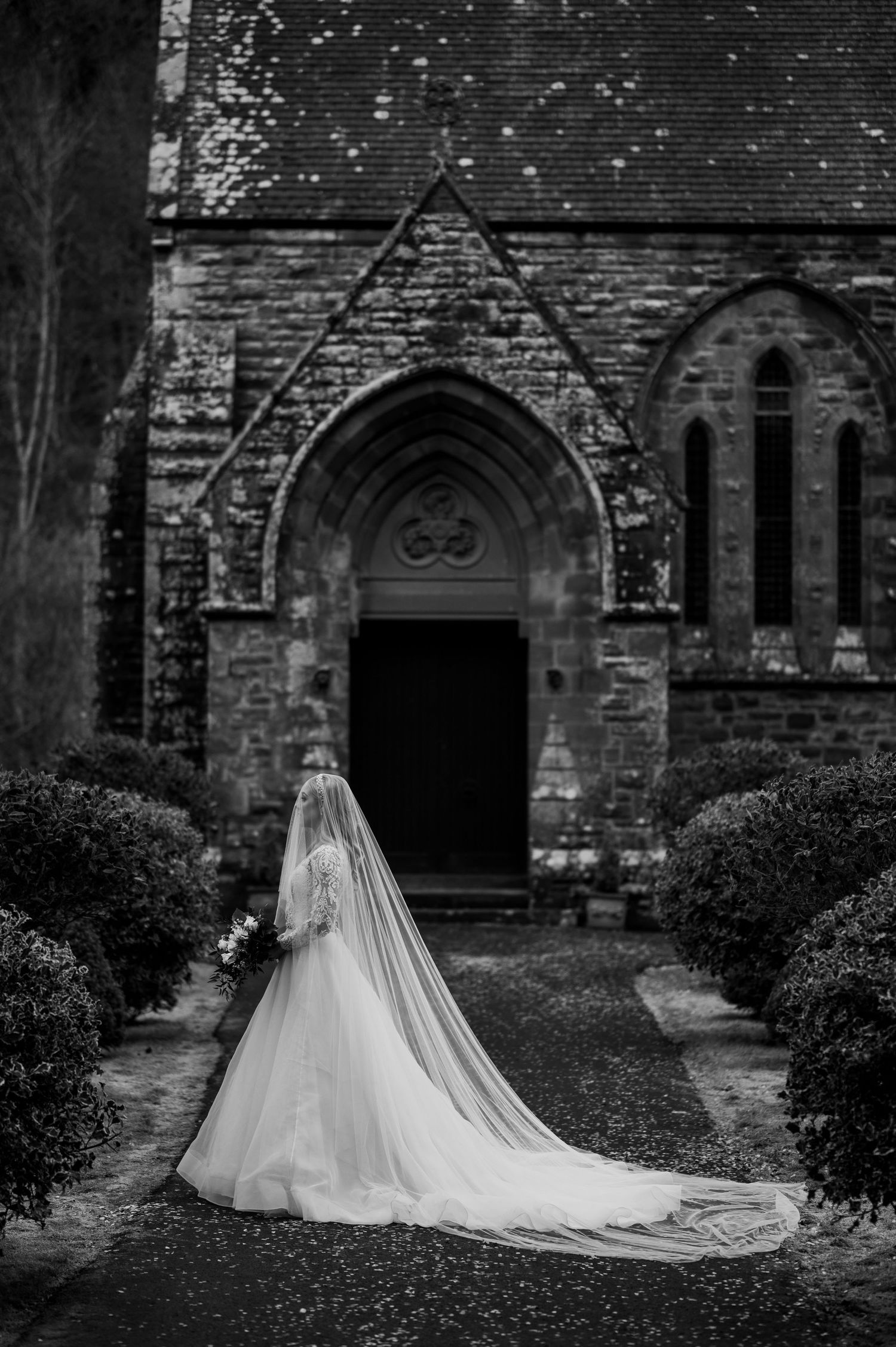 Aberdeen Wedding Photography. A flowing white wedding dress with a long train photographed in front of a gothic stone church entrance.