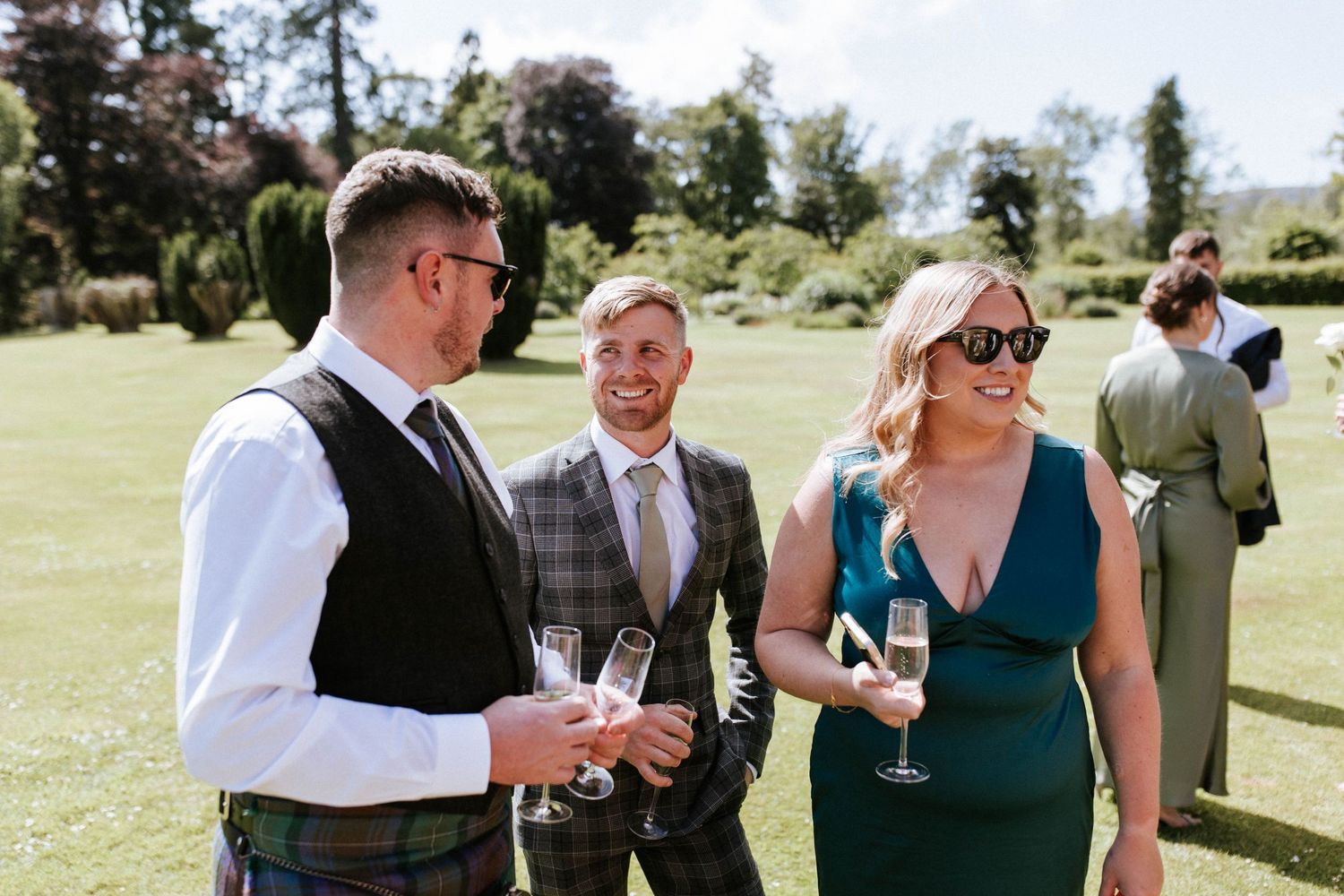 Wedding guests in formal attire socializing with drinks on a sunny lawn.