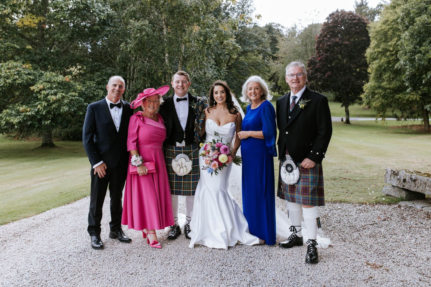 Small wedding group wearing mix of formal attire and kilts poses outdoors on garden path.