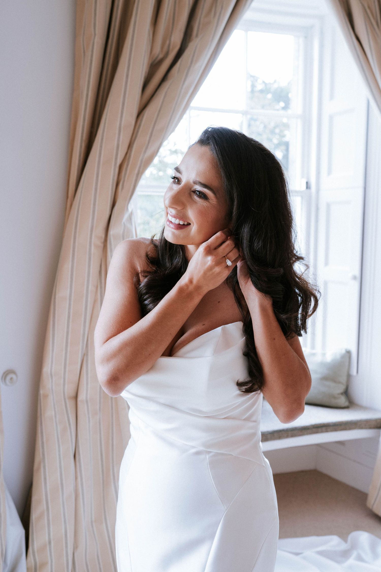 A bride in a white dress touches her hair while standing by a window with beige curtains.