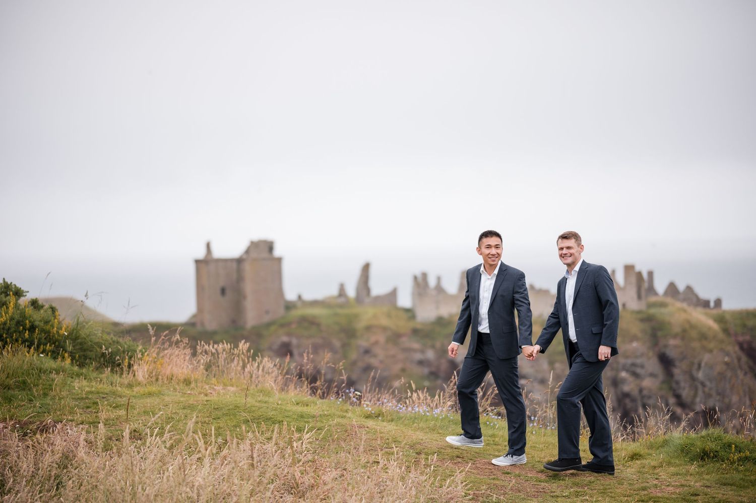 Same Sex Wedding Photography. Two people walk together across a windswept coastal meadow with ancient castle ruins on the horizon.