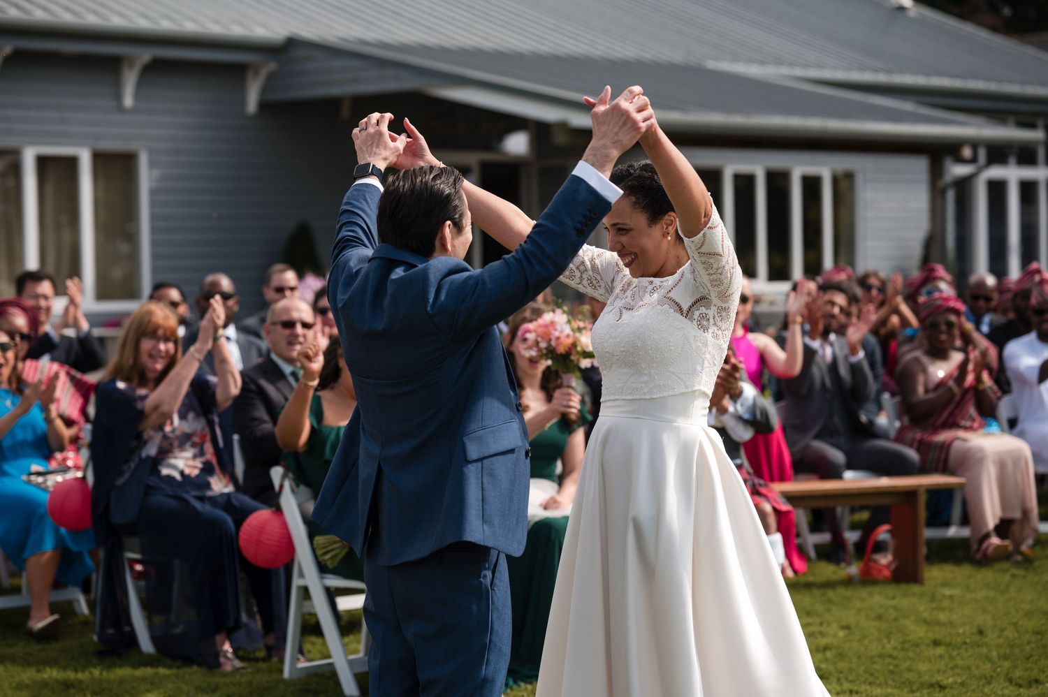 Wedding celebration on a lawn with guests seated in white chairs near a grey building.