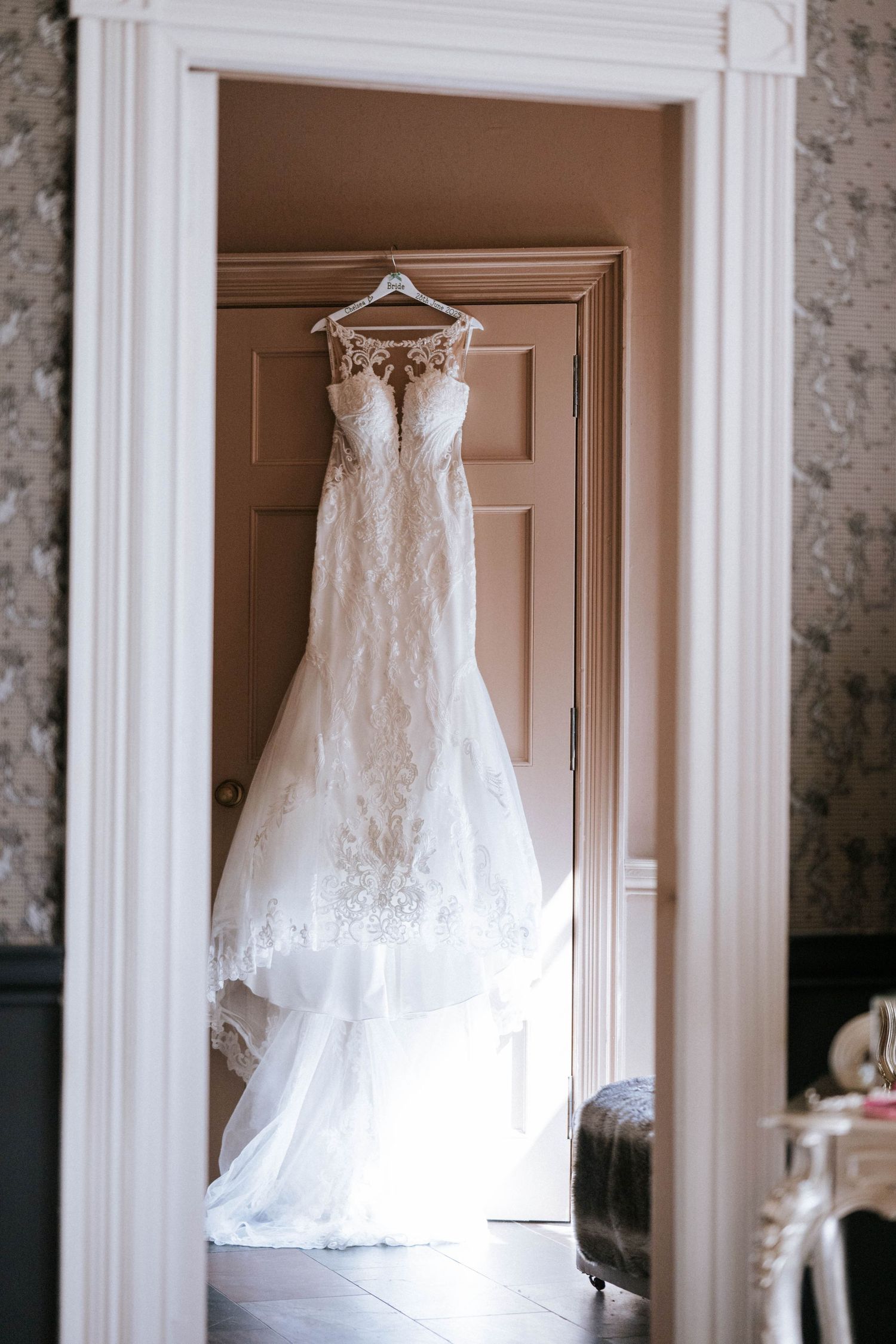 Elegant white lace wedding dress hanging in a doorway against a neutral-colored wall.