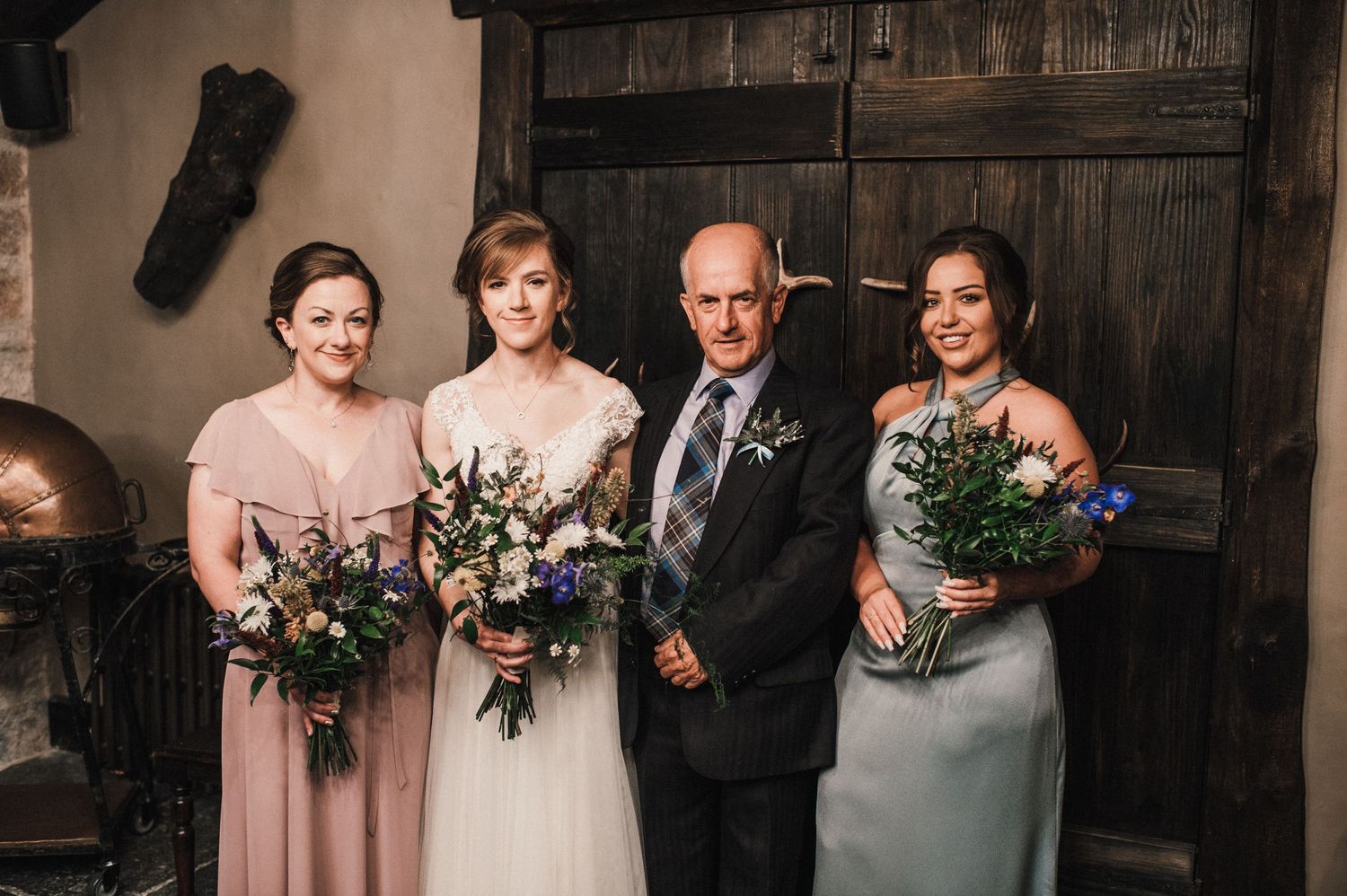 Wedding party members pose together holding floral bouquets against rustic wooden doors.