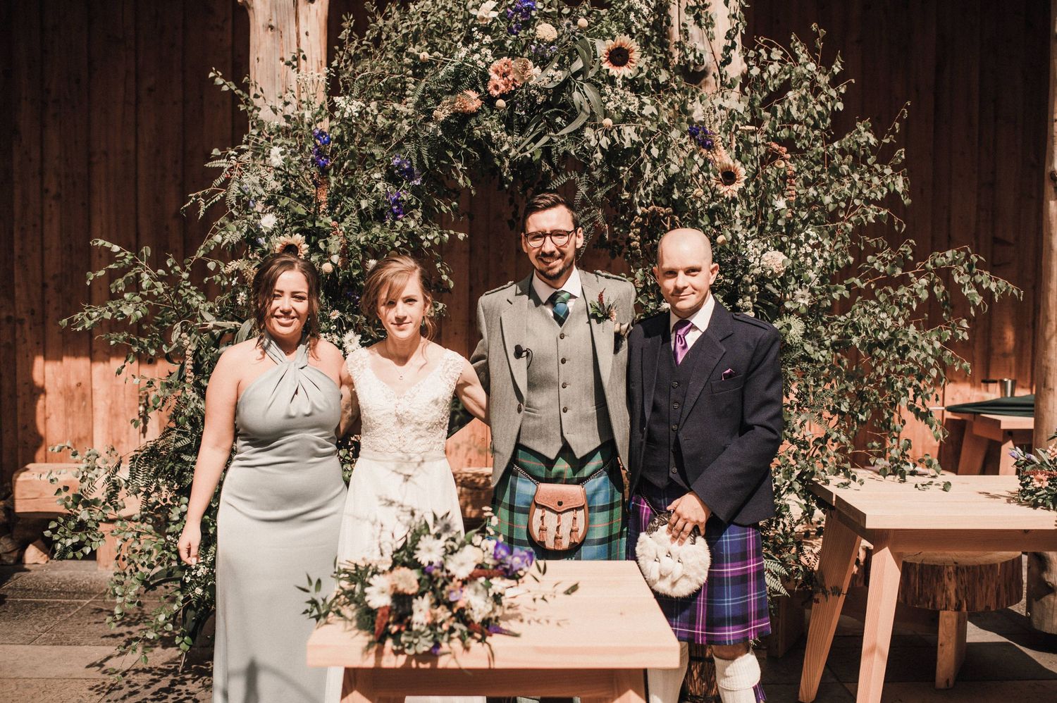 Wedding party poses together under a floral arch with traditional Scottish kilts and formal attire.