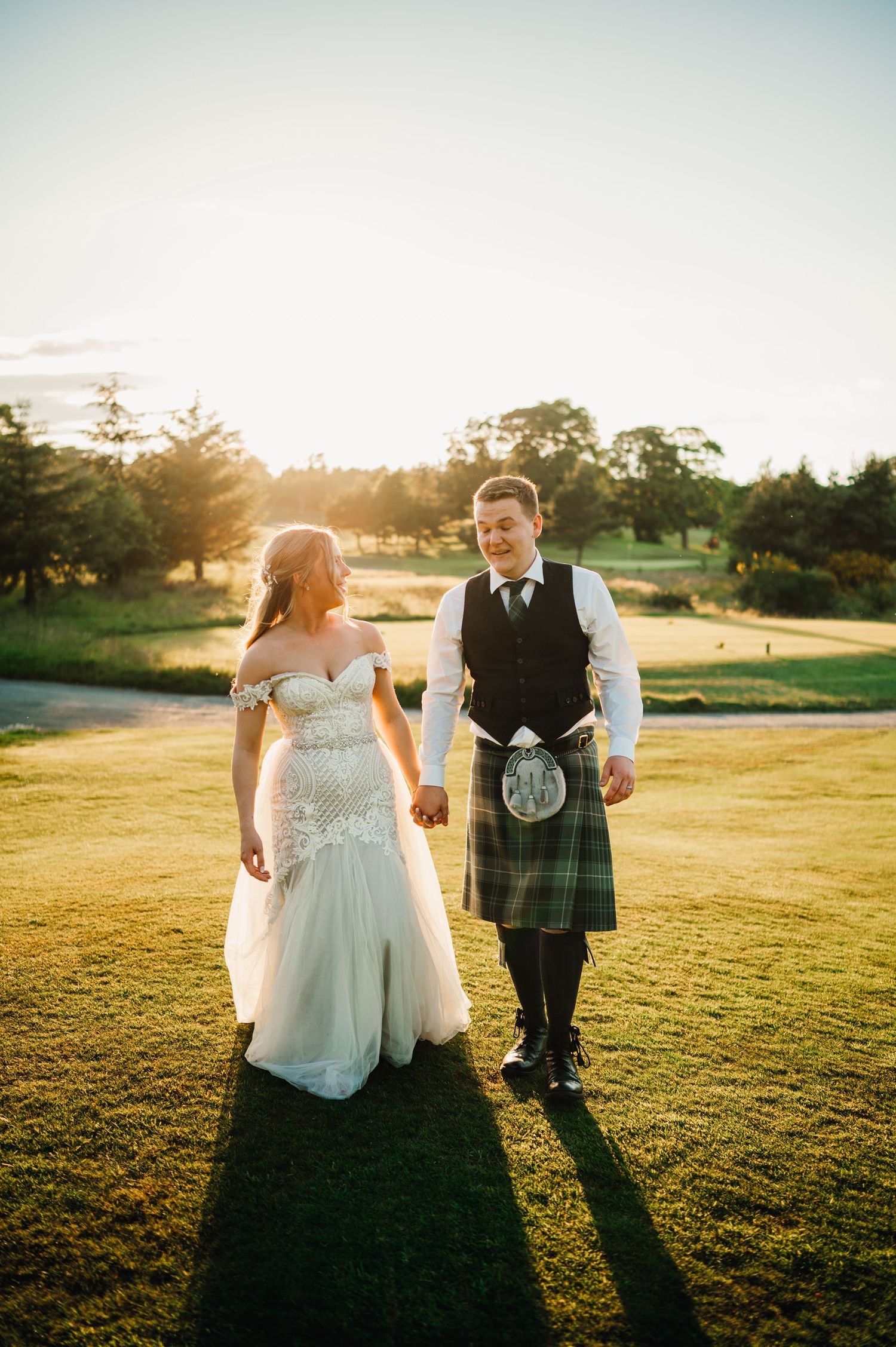 A couple walking on a golf course during sunset with the groom wearing a traditional Scottish kilt.