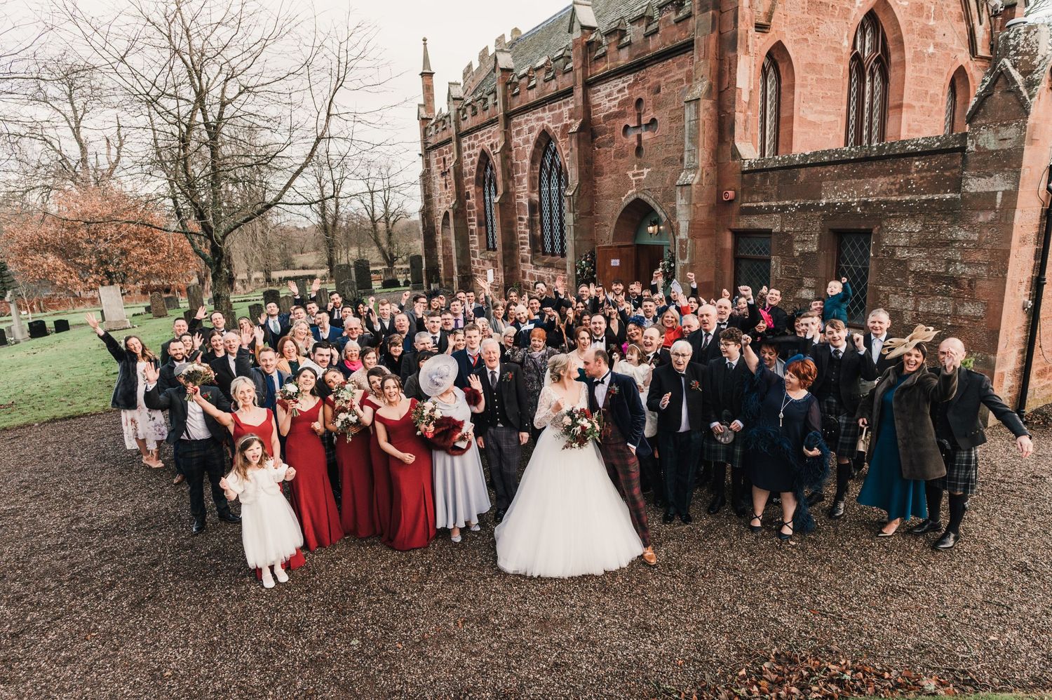 Large wedding party group photo gathered outside historic stone church with archway entrance.