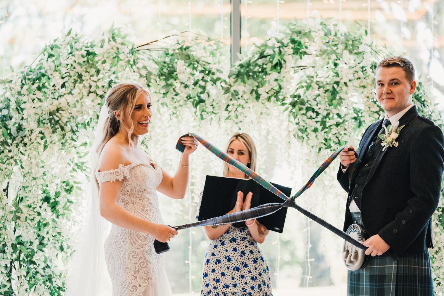 A wedding ceremony under a floral arch with traditional Scottish attire and decorative elements.