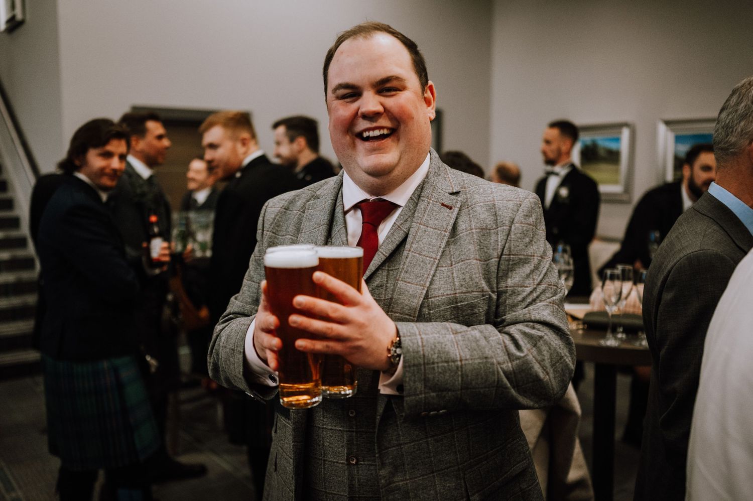 A guest in a gray suit and red tie enjoys a beer at an indoor social gathering.