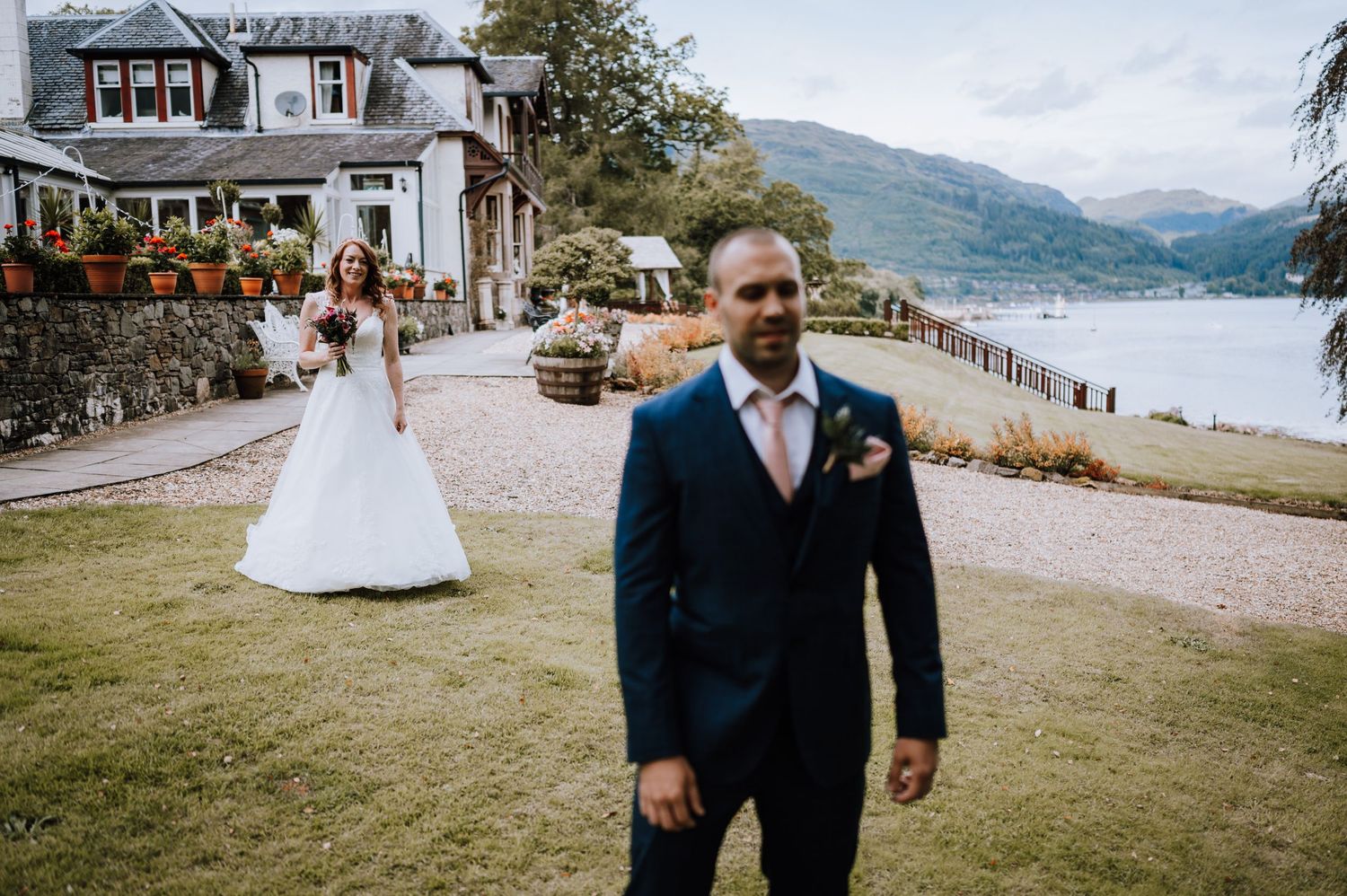 Bride and groom pose on lawn near lake and building with mountain backdrop.