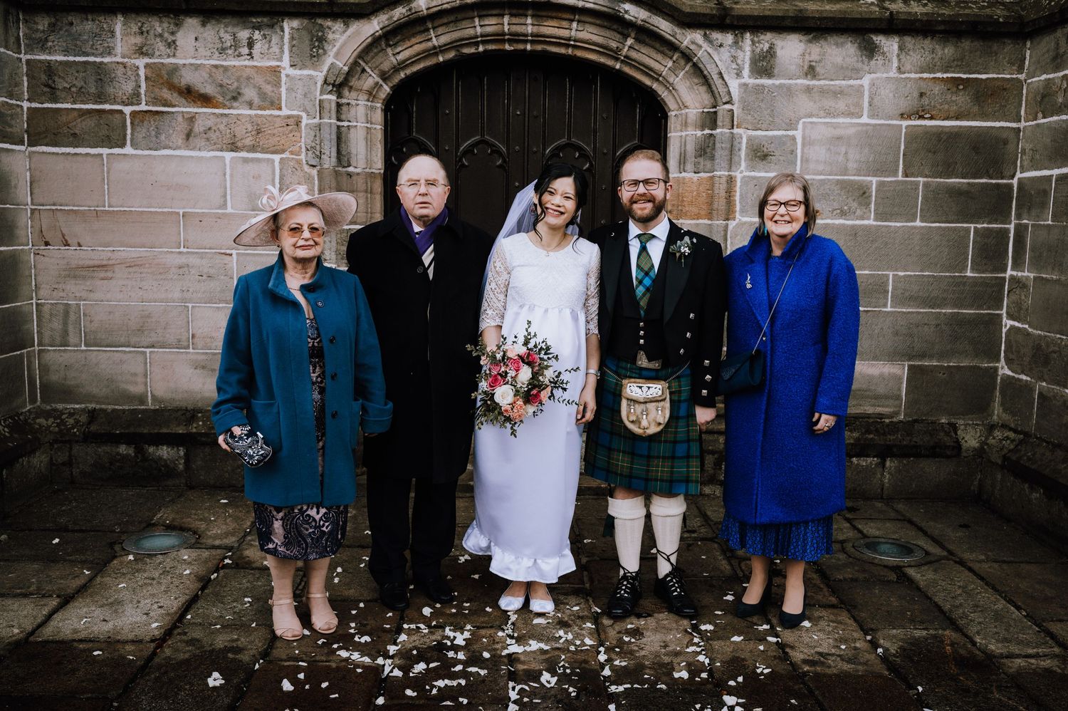 Family portrait in front of stone church with Scottish wedding attire.