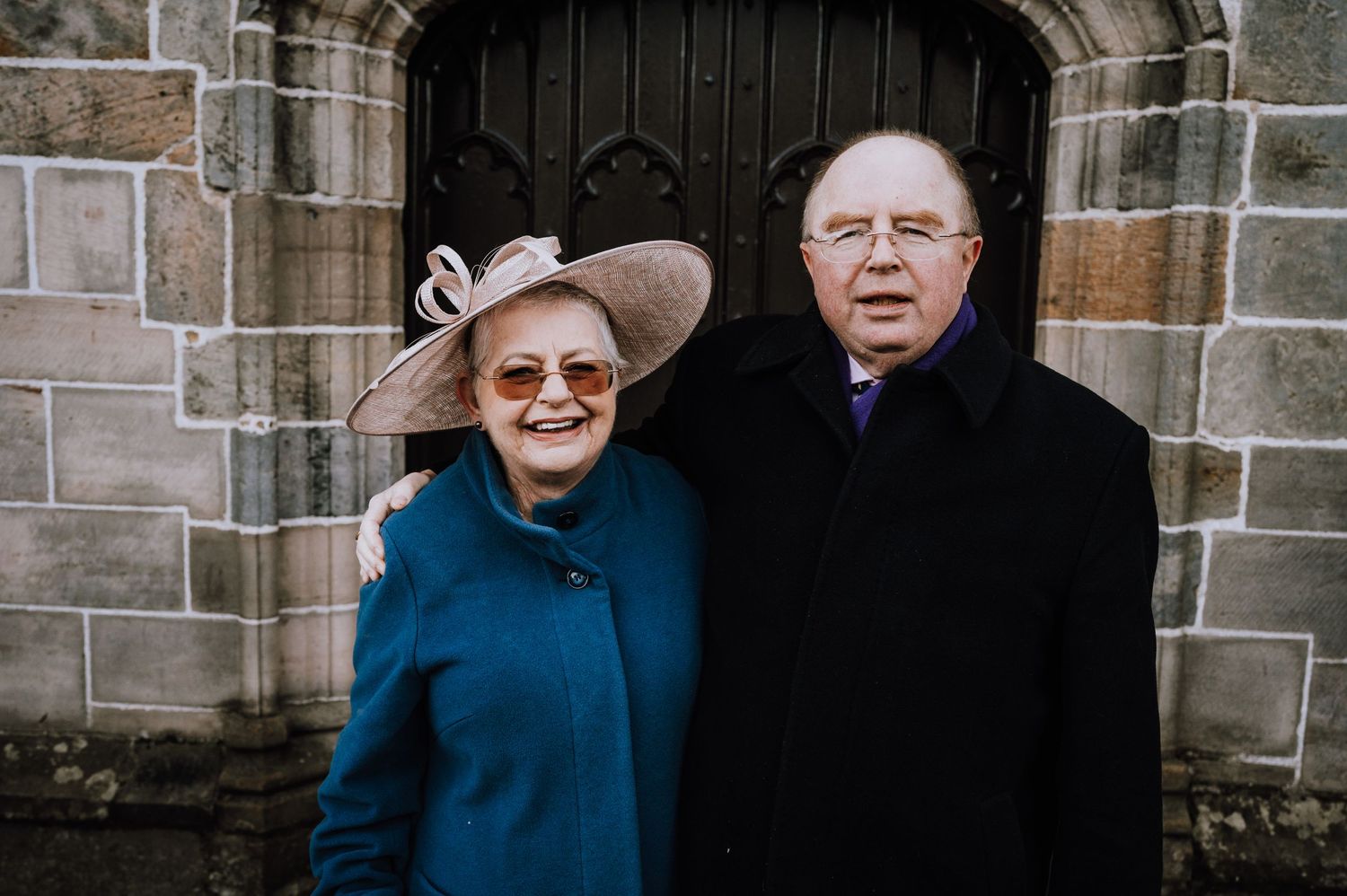 An elderly couple stands in front of a stone church archway, with the woman wearing a blue coat and wide-brimmed hat.