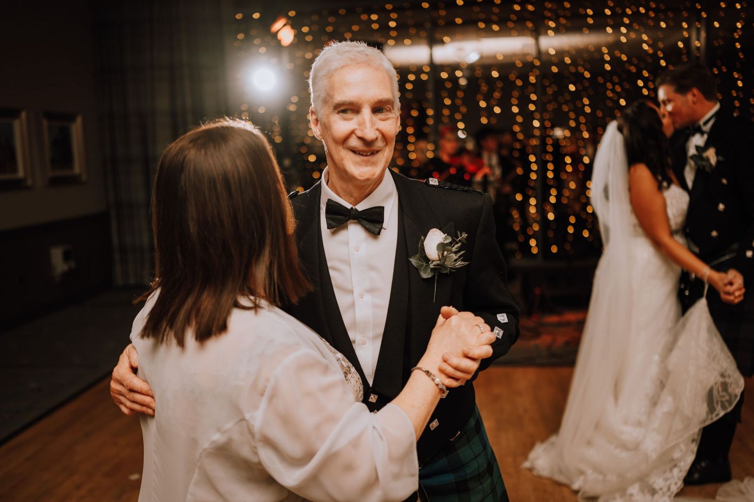 Wedding guests dancing on wooden floor with string lights creating warm ambiance.