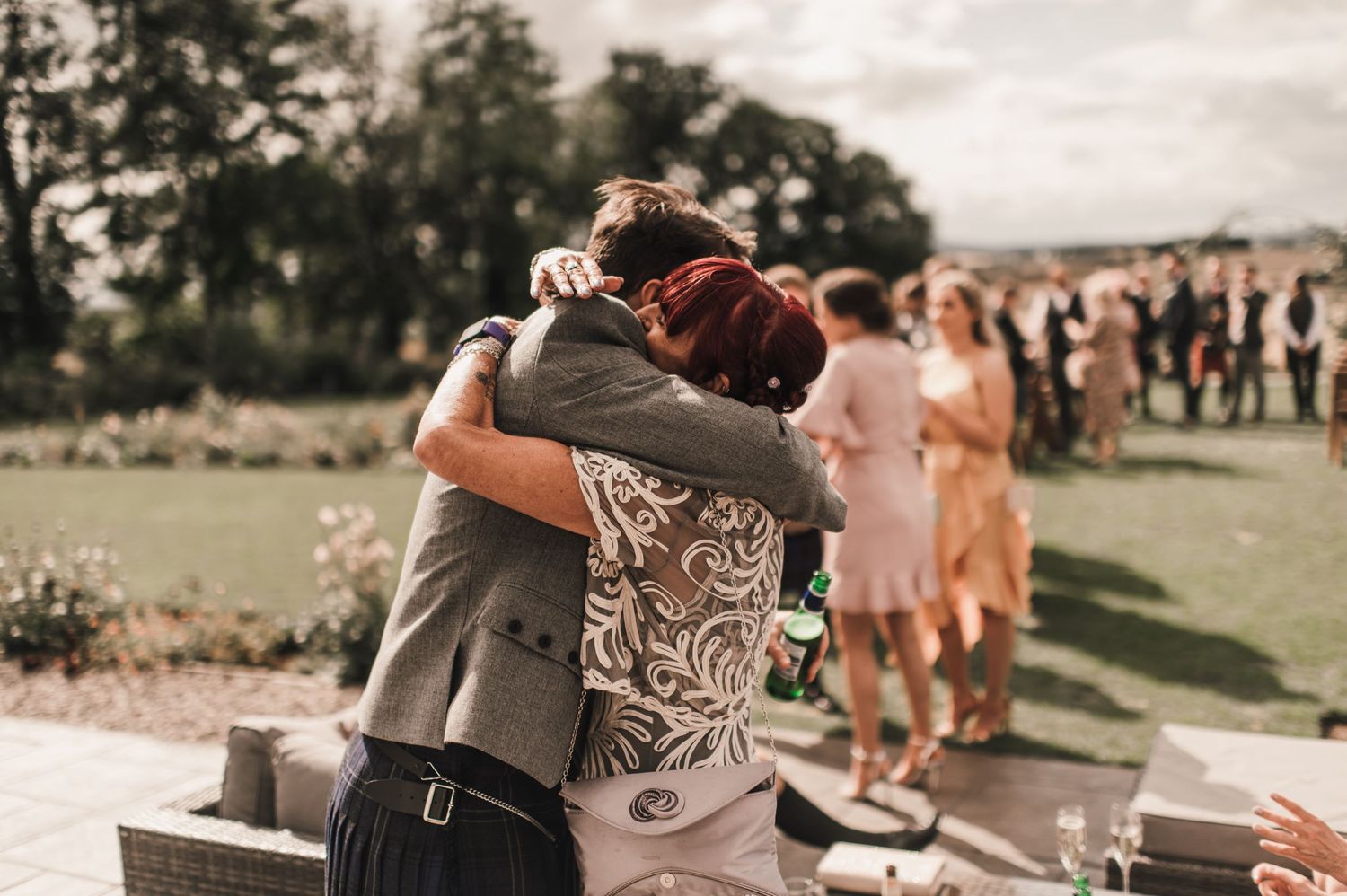 Wedding guests embrace during an outdoor celebration with others in the background.