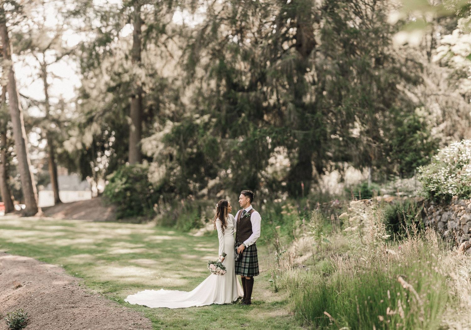 Wide landscape shot of newlyweds in Scottish attire standing together on forest path.
