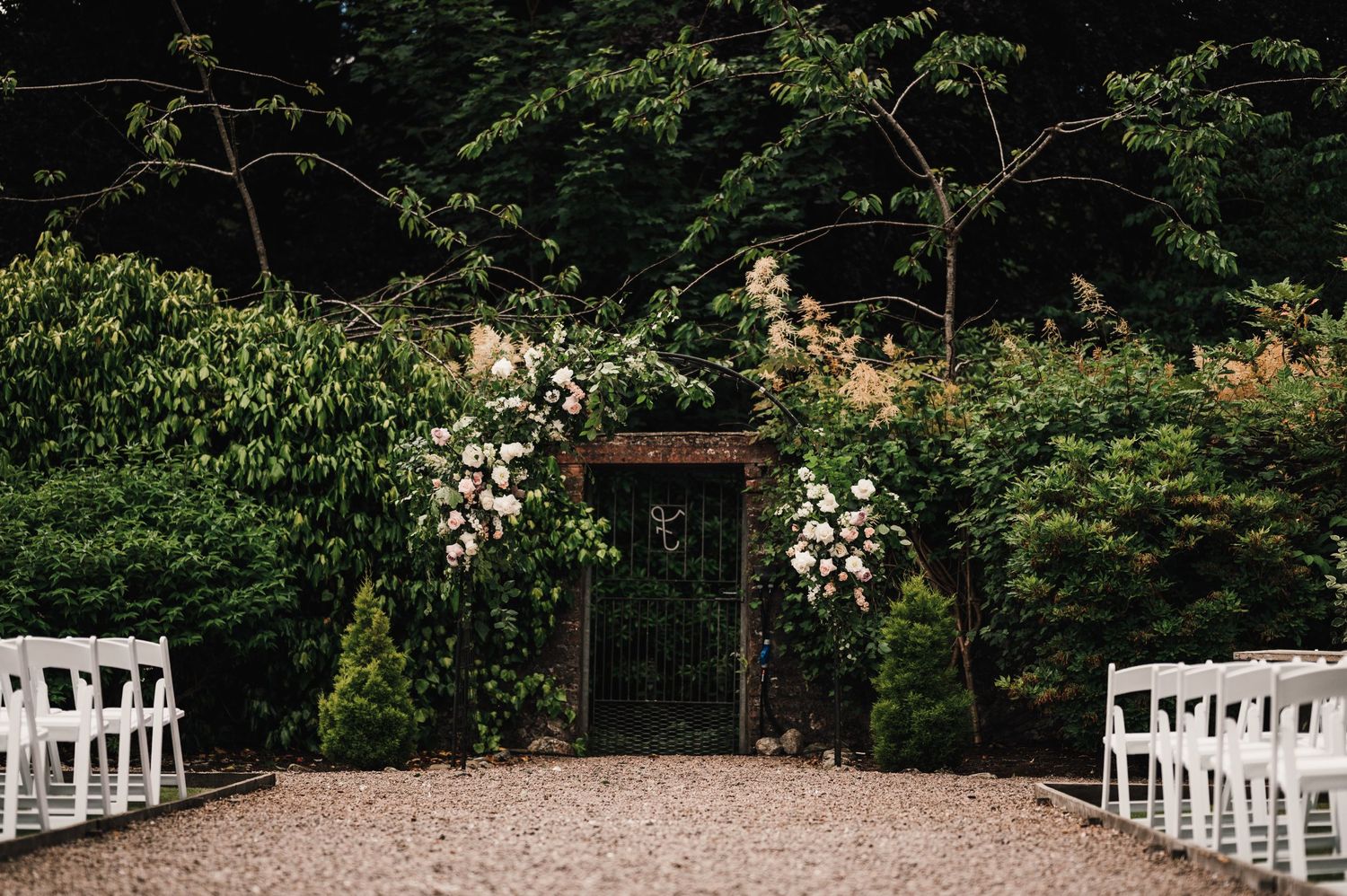 Garden wedding arch decorated with white roses and greenery, with white chairs lined up for ceremony.