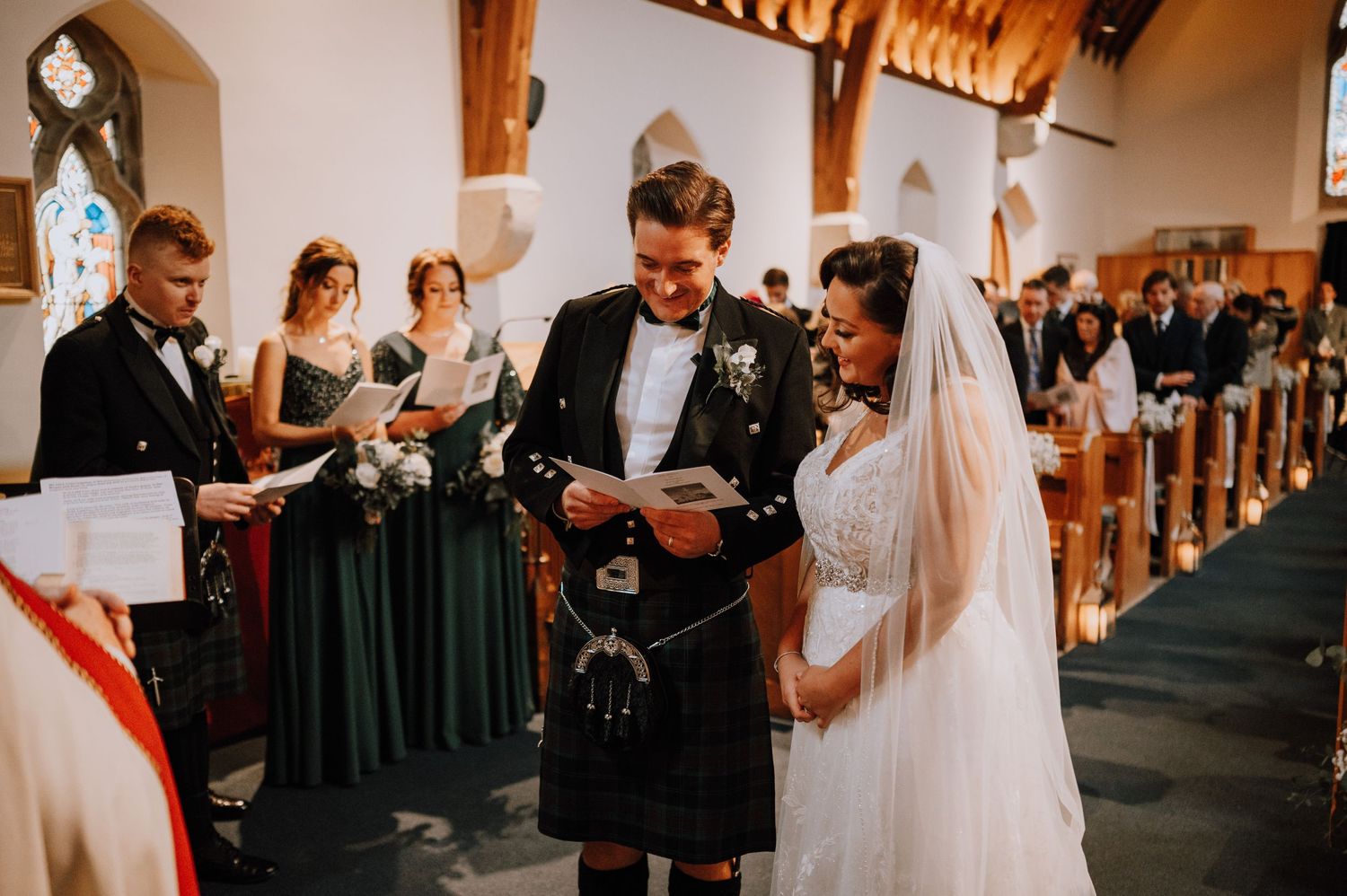 A wedding ceremony in a church with bridesmaids in dark green dresses and Scottish attire.