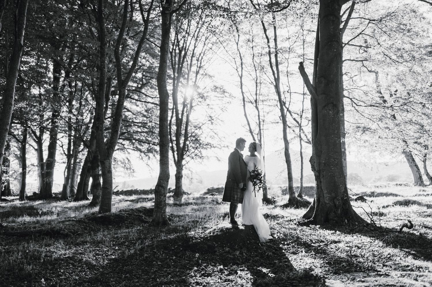 A romantic black and white wedding photo showing a couple embracing among tall trees in a forest setting.